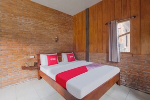 Una habitación con pared de ladrillo, paneles de madera, cama con ropa de cama blanca y detalles en rojo, una pequeña estantería con una cesta y una ventana con cortinas.