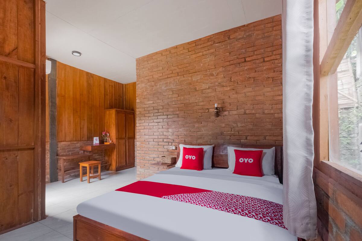 A bedroom with a brick wall, wooden furniture, a bed with red and white bedding, and a window with a view.