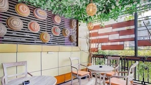 Un café avec des tables et des chaises rondes, un mur décoré de paniers tressés et une vue sur la verdure grâce à de grandes fenêtres.