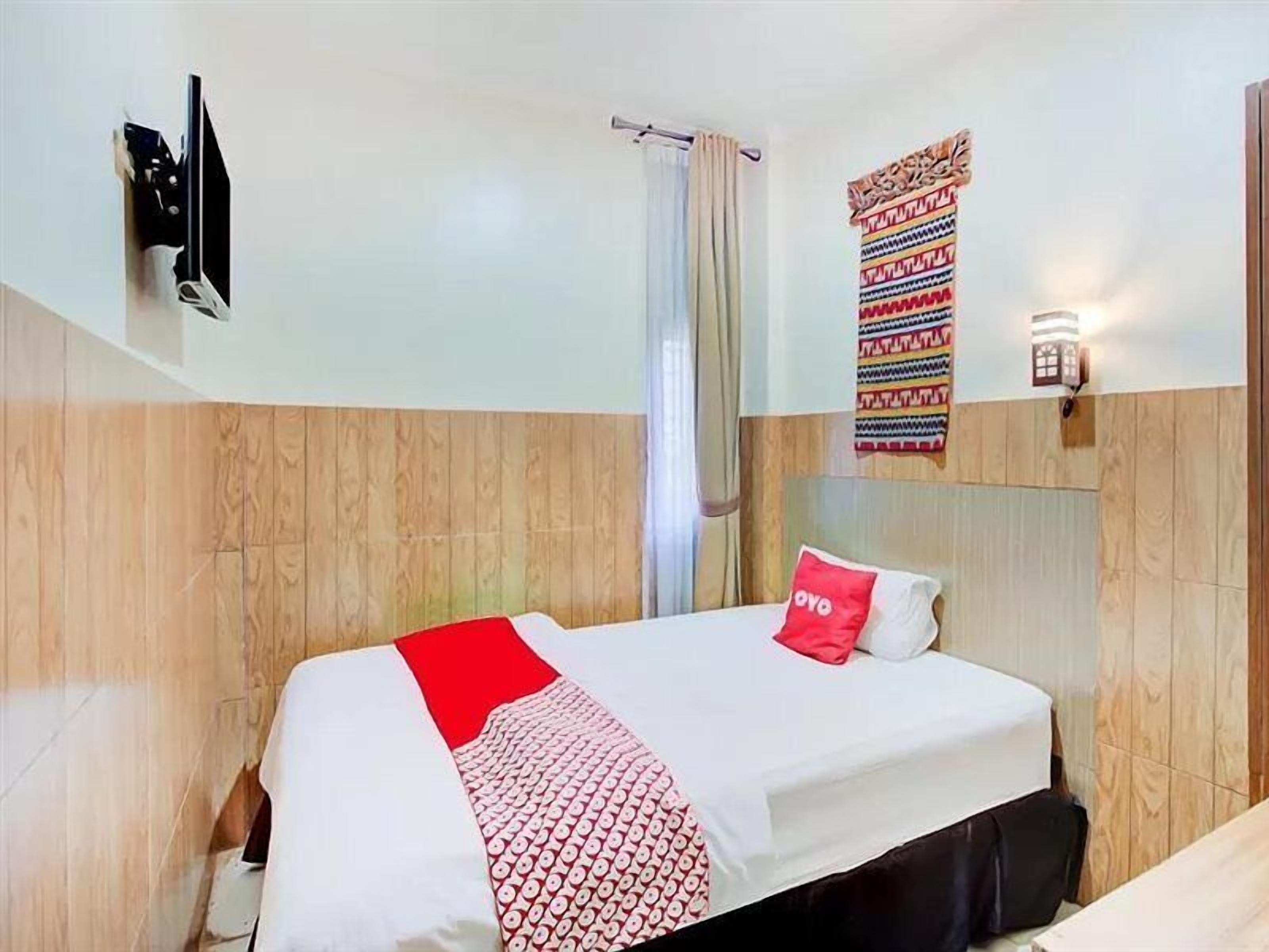 A hotel room with a bed, a TV mounted on the wall, a window with curtains, and a wall-mounted light fixture.
