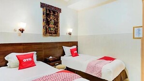 Une chambre d’hôtel avec deux lits, chacun agrémenté d’un oreiller rouge orné du logo « OYO » et d’une couvre-lit à motifs. Un tapis décoratif orne le mur, et une petite table de chevet avec une lampe se trouve à côté du lit.