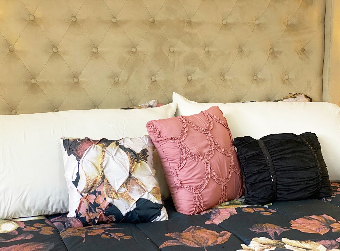 Ein Bett mit einer gepolsterten Kopfteile, einer zudeckenden Bettdecke mit Blumenmuster und drei Kissen in unterschiedlichen Designs.