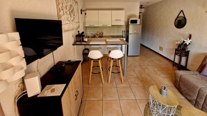 Un espacio habitable compacto con kitchenette, un televisor montado en la pared, una pequeña zona de comedor con taburetes y un sofá.