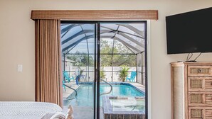 Habitación de hotel con un amplio ventanal corredero que da a una zona de piscina, una cama con manta blanca y un televisor montado en la pared.
