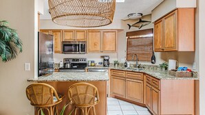 Cocina con armarios de madera, microondas, estufa y pileta. Hay dos sillas de mimbre y una mesa con mantel azul.