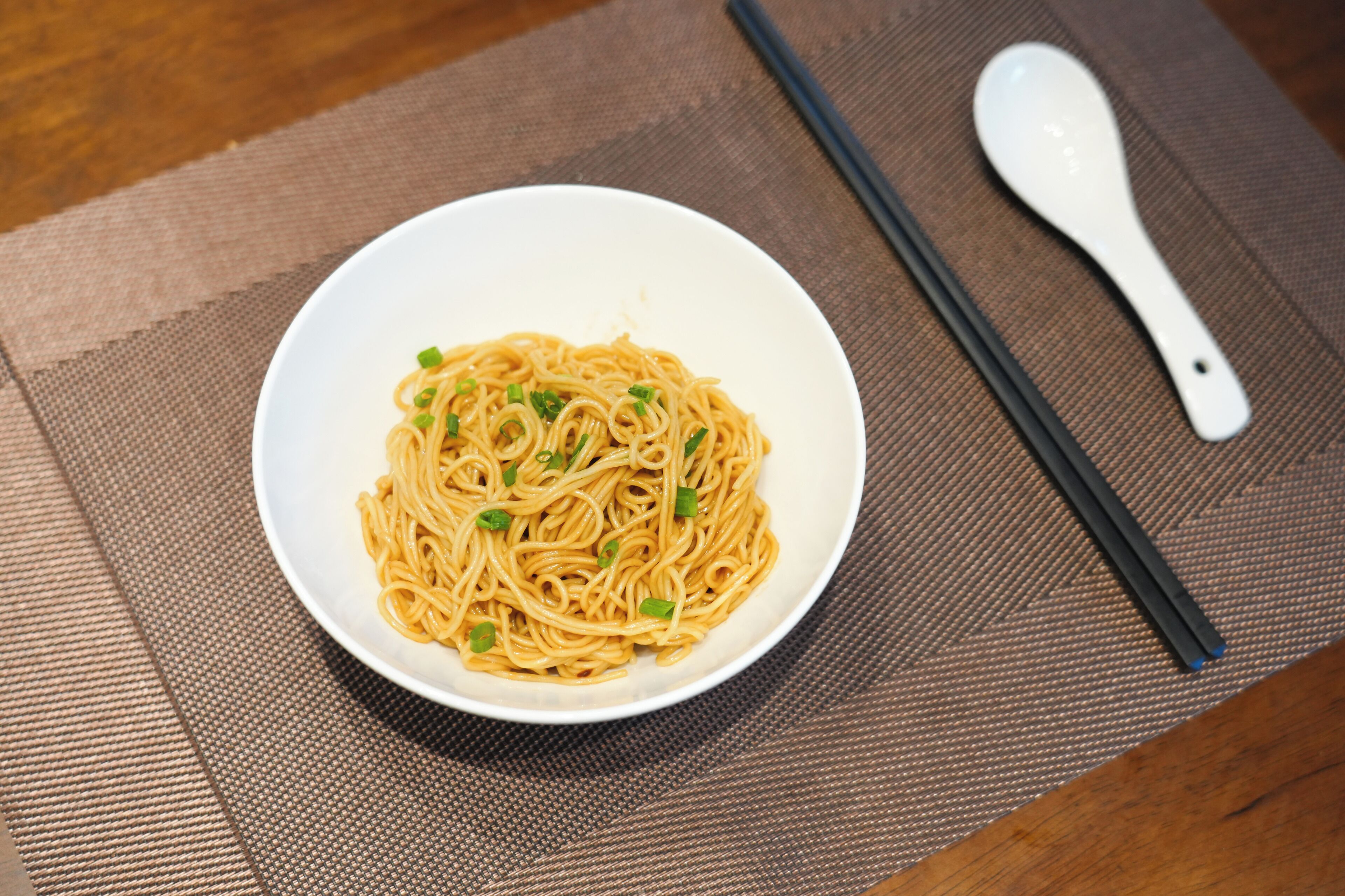 A bowl of noodles on a textured mat with chopsticks and a spoon beside it.
