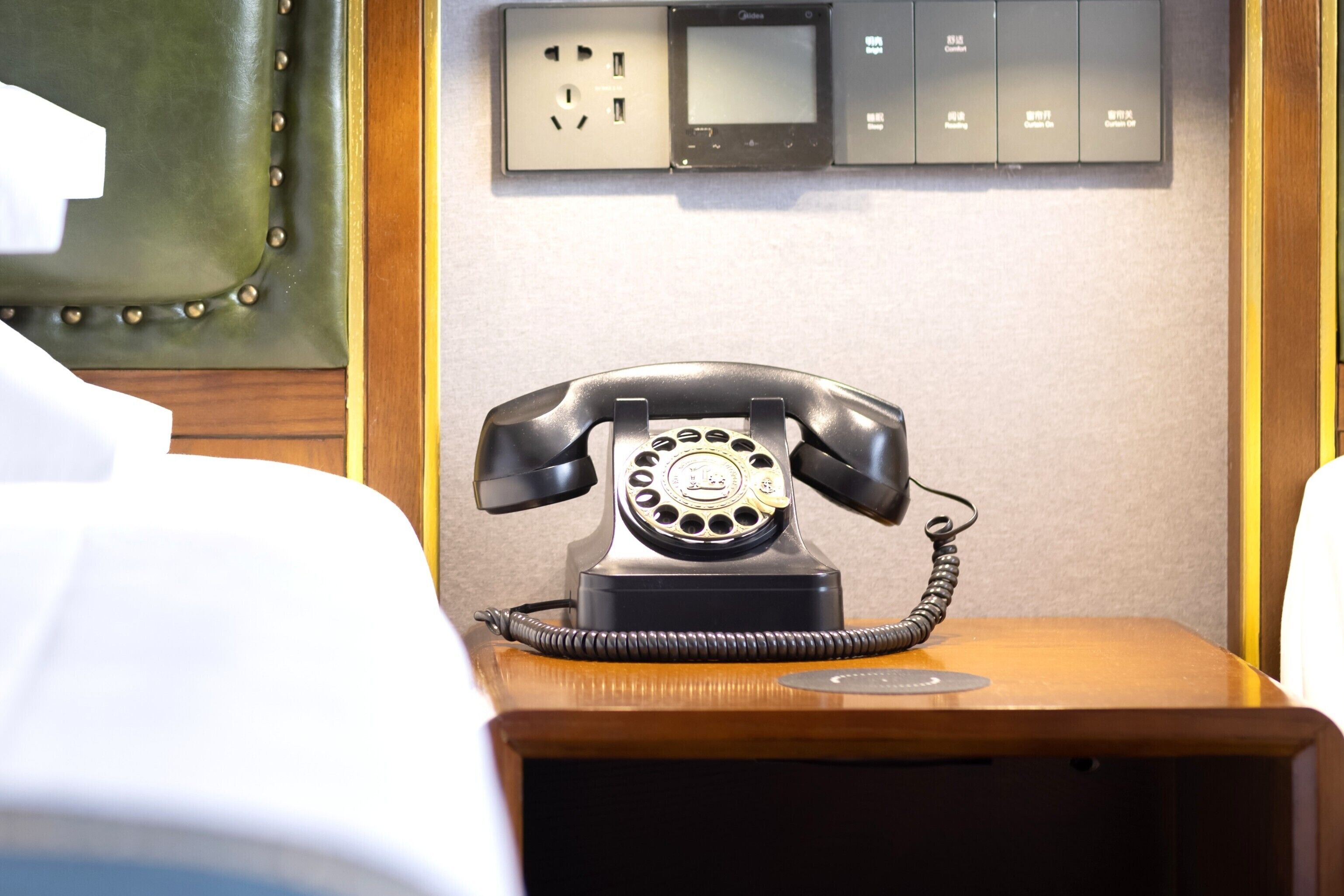 A hotel room with a vintage rotary phone on a wooden nightstand, a bed with white linens, and a wall-mounted electrical panel with a digital display.