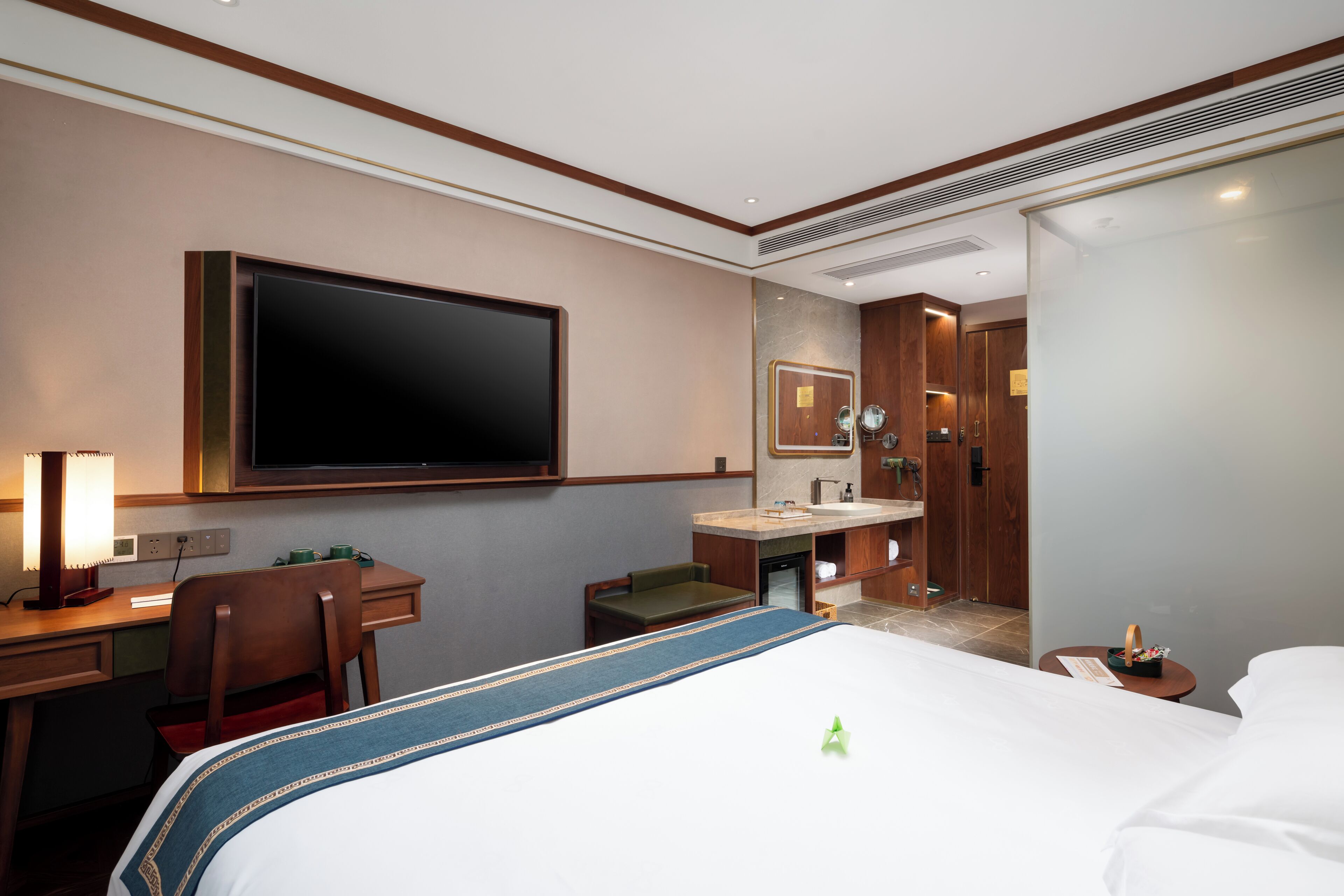 A hotel room with a large bed, a desk with a chair, a television, a bathroom with a sink and mirror, and a small table with a bottle.