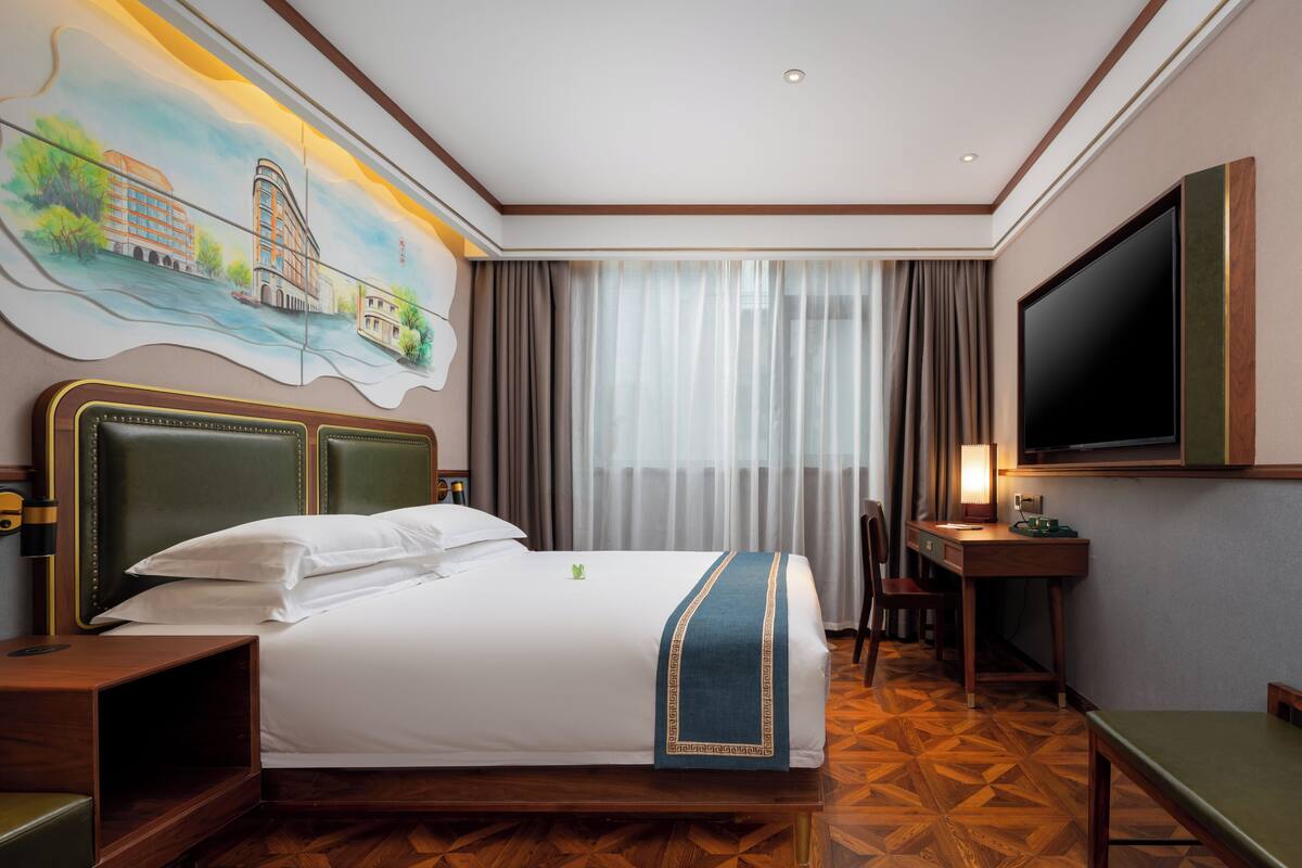 Ein Hotelzimmer mit einem großen Bett, einem Schreibtisch, einem Sessel, einem Fernseher und einem Bild an der Wand.