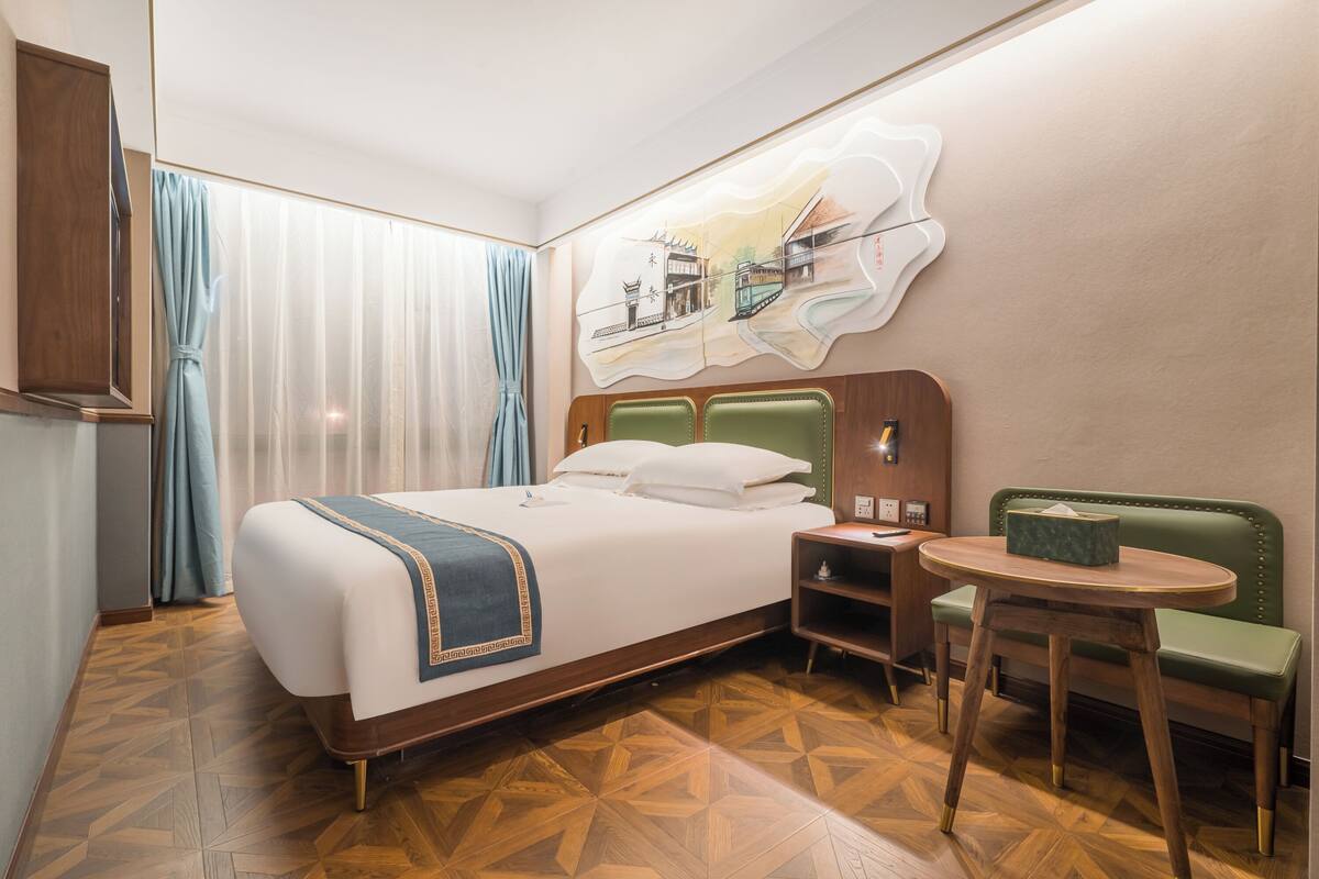 Ein Hotelzimmer mit einem Bett, einer grünen Polsterbank, einem kleinen runden Tisch, einem Nachttisch und einem Wandbild.