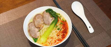 A bowl of noodle soup with meatballs and vegetables on a woven placemat.