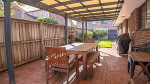 A covered patio area with a wooden table and chairs, a barbecue grill, and a brick floor.