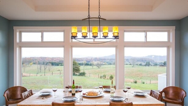 Uma sala de jantar com uma grande mesa de madeira posta para seis pessoas, grandes janelas com vista para uma paisagem rural e um lustre pendurado acima.