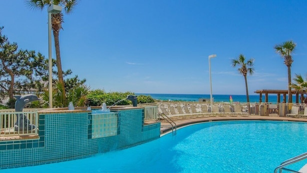 A clear blue pool with a tiled edge, surrounded by palm trees and a view of the ocean.