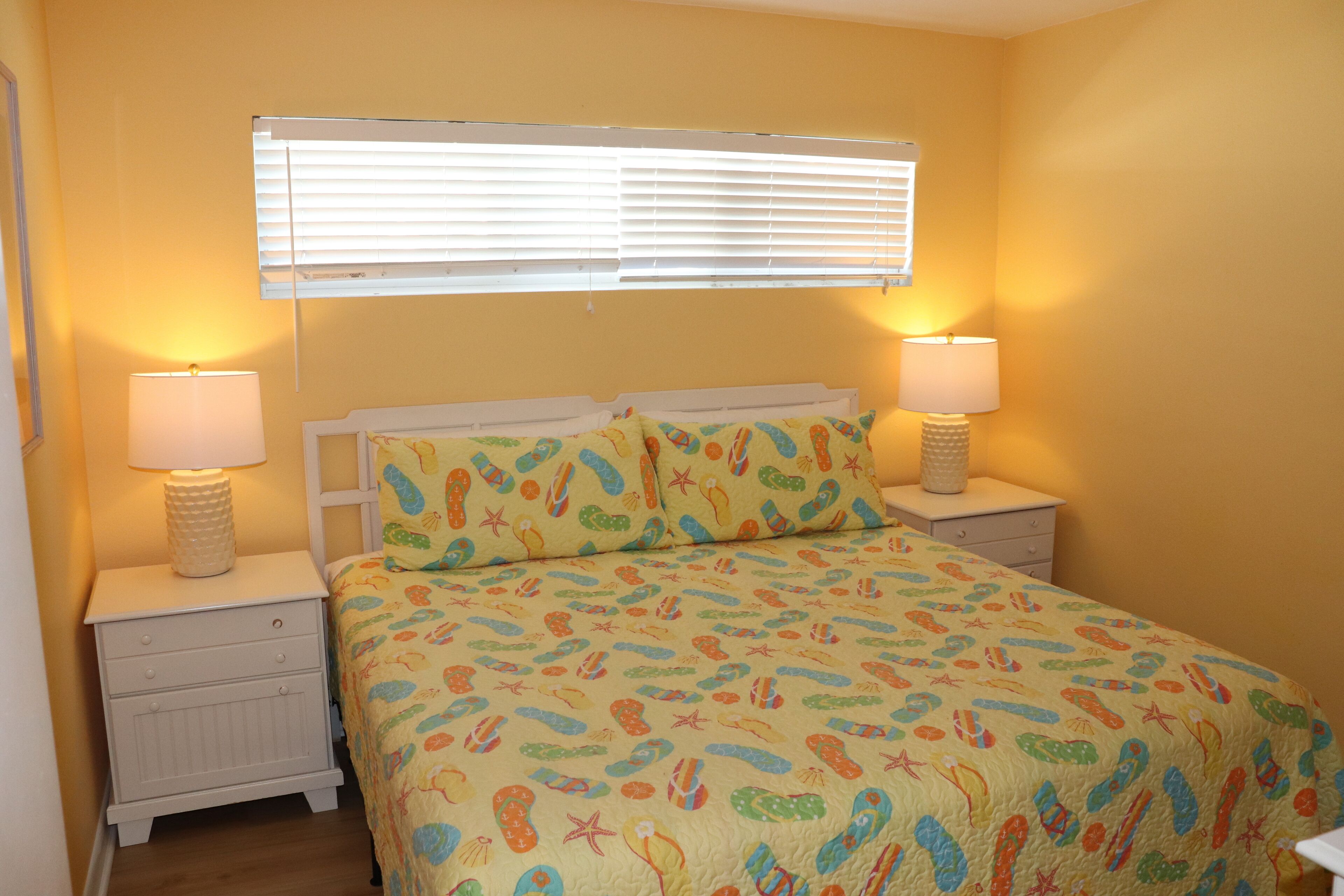 A bedroom with a bed, two bedside tables, a window with blinds, and a wall with a yellow hue.