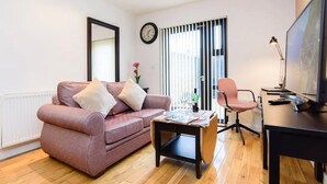 A modern living room with a pink sofa, a coffee table, a desk with a computer, and a large window with blinds. - 15% Off Work Week Rates | 1Bd Apt w/Pvt Terrace (Cardiff)