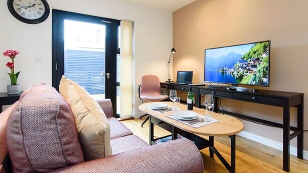 A modern living room with a sofa, a dining table set for two, a TV on a stand, and a clock on the wall. - 15% Off Work Week Rates | 1Bd Apt w/Pvt Terrace (Cardiff)