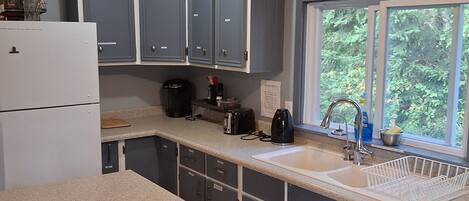 A kitchen with grey cabinets, a white countertop, a sink, a coffee maker, and a window with a view of greenery.