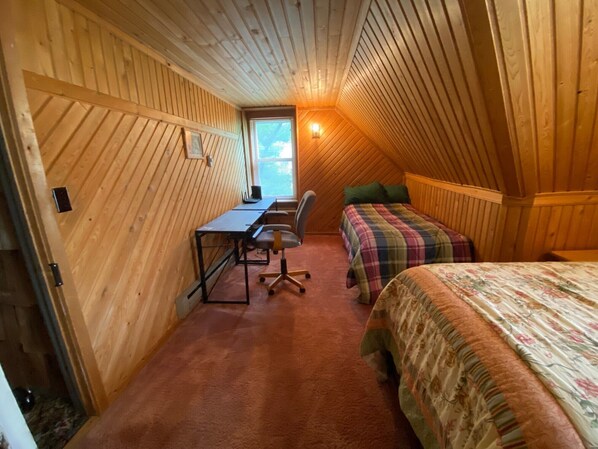 Une chambre confortable située dans les combles, avec un plafond en pente en bois, un bureau avec une chaise, un lit recouvert d’une couvre-lit à motifs floraux et une fenêtre avec des stores.
