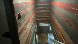 A narrow staircase with wooden walls painted in horizontal stripes of red, white, and gray. A light fixture hangs from the ceiling, and the staircase leads to a landing with a wooden railing. - House in Bessemer - 3 BR/1.5 Bathrooms (Bessemer)
