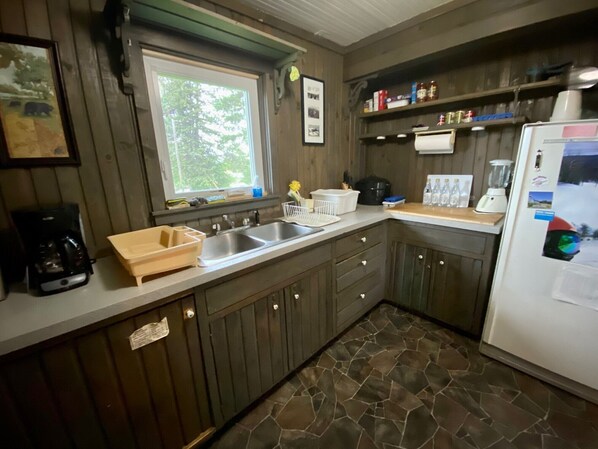A kitchen with wooden cabinets, a double sink, a coffee maker, a refrigerator, and a window with a view of trees.