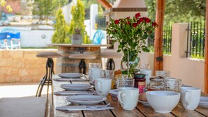 A wooden table set for breakfast with white plates, cups, and bowls, a vase with red flowers, and glass jars of jam. - Nautilus Villa with private pool (Hersonissos)