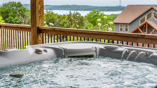 Un bain à remous avec une terrasse en bois, entouré de verdure et avec une maison en arrière-plan.