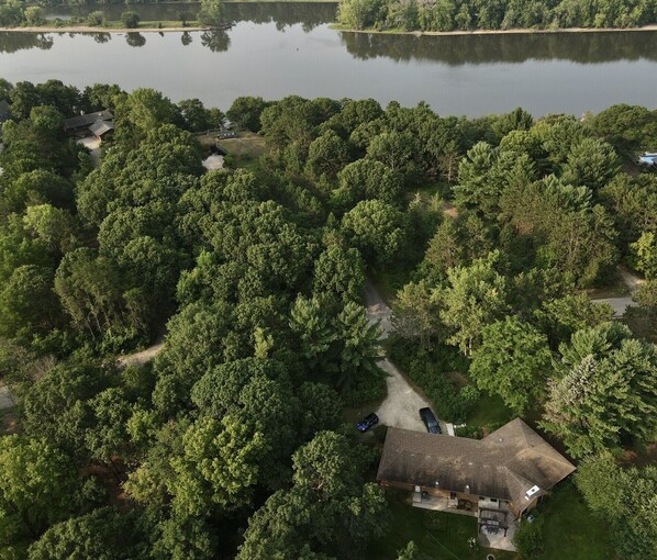 Vista aérea de una zona residencial con una casa rodeada de árboles frondosos, un camino de entrada visible y una masa de agua al fondo.