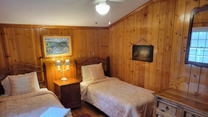 Una habitación con paneles de madera, dos camas, una mesita de noche con lámparas, un aparador con cajones y un cuadro en la pared.