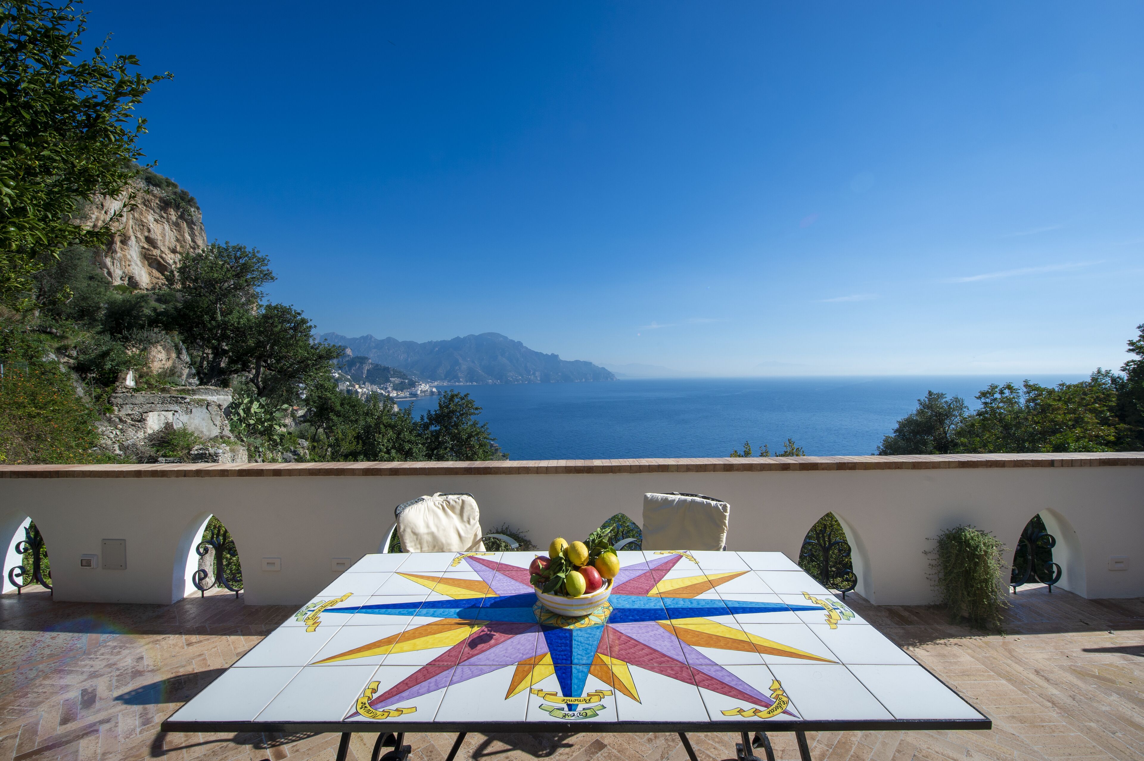 A table with a colorful tiled top, a bowl of fruit, and a view of the sea and mountains.
