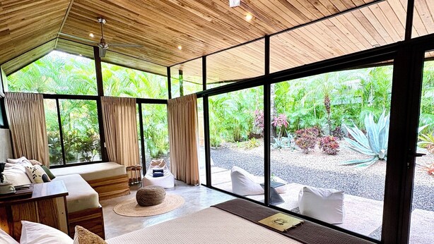 Un dormitorio amplio con una cama grande, un ventilador de techo y vistas a un jardín exuberante a través de amplios ventanales.