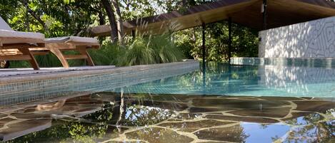 A clear swimming pool with tiled patterns, surrounded by lounge chairs and a covered seating area.