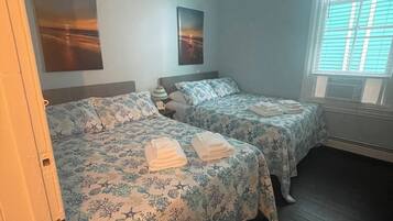 Une chambre avec deux lits, chacun agrémenté de draps et de serviettes à motifs floraux. On y trouve deux tableaux accrochés au mur, un luminaire au plafond et une fenêtre avec des stores.