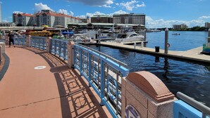 A waterfront promenade with a blue metal railing, a paved walkway, and a marina with boats docked.
