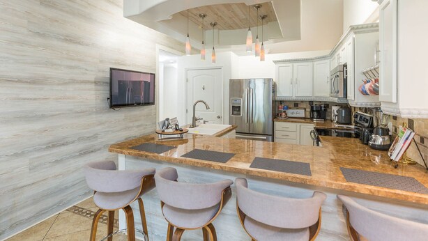 A modern kitchen with a marble countertop, stainless steel appliances, and a flat-screen TV mounted on the wall.