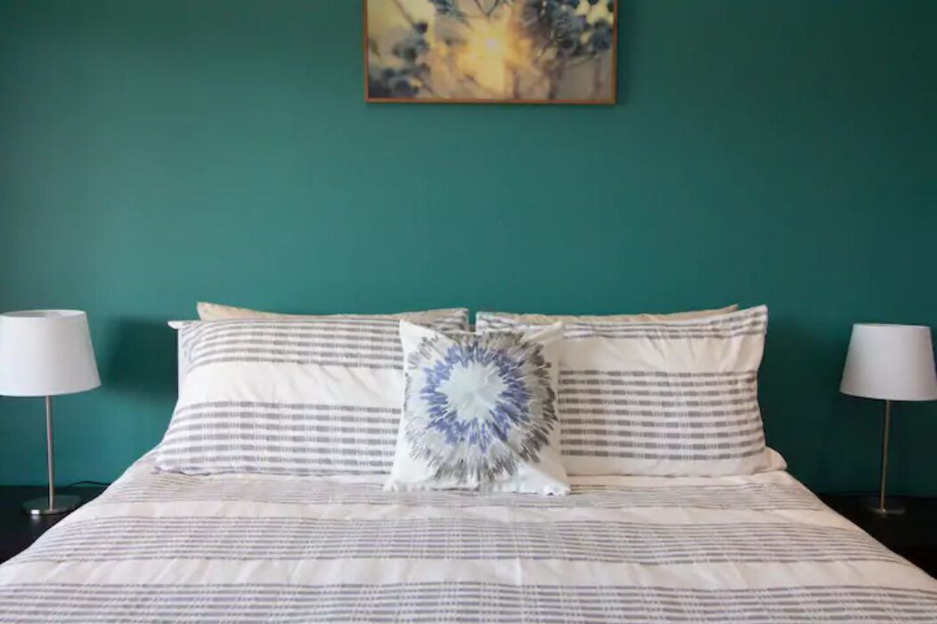 A neatly made bed with striped bedding and a decorative pillow, flanked by two bedside lamps, against a teal wall with a framed artwork above.