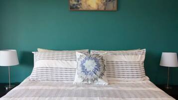 A neatly made bed with striped bedding and a decorative pillow, flanked by two bedside lamps, against a teal wall with a framed artwork above.