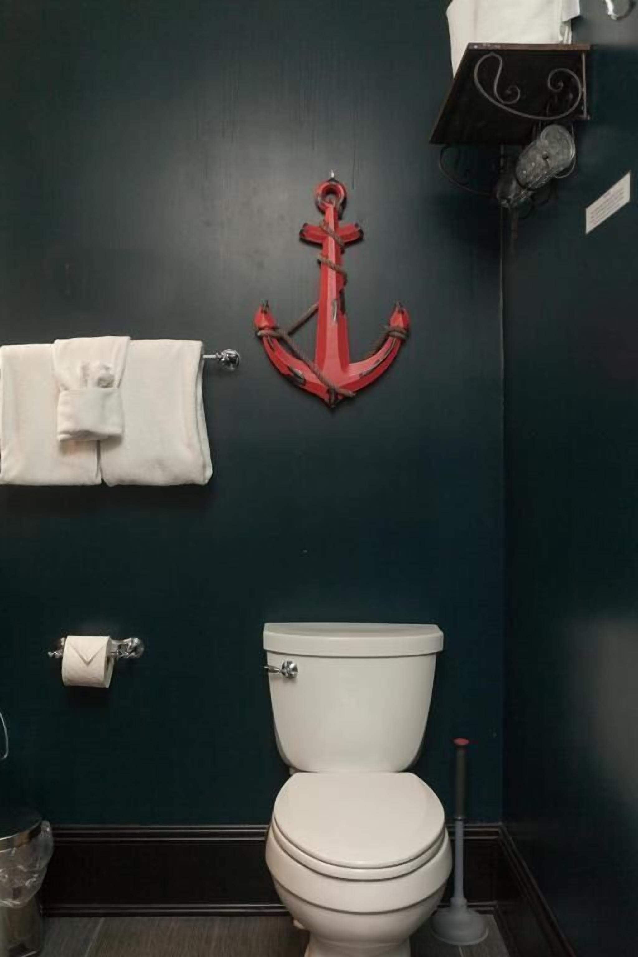 Een badkamer met een donkergroene wand, een wit toilet, een rode decoratie van een anker, een handdoekhouder met een opgevouwen handdoek, en een toiletpapierhouder.
