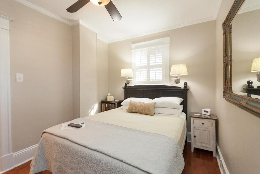 Un dormitorio ordenado con cama, lámparas de noche, mesita de noche, espejo y ventilador de techo.