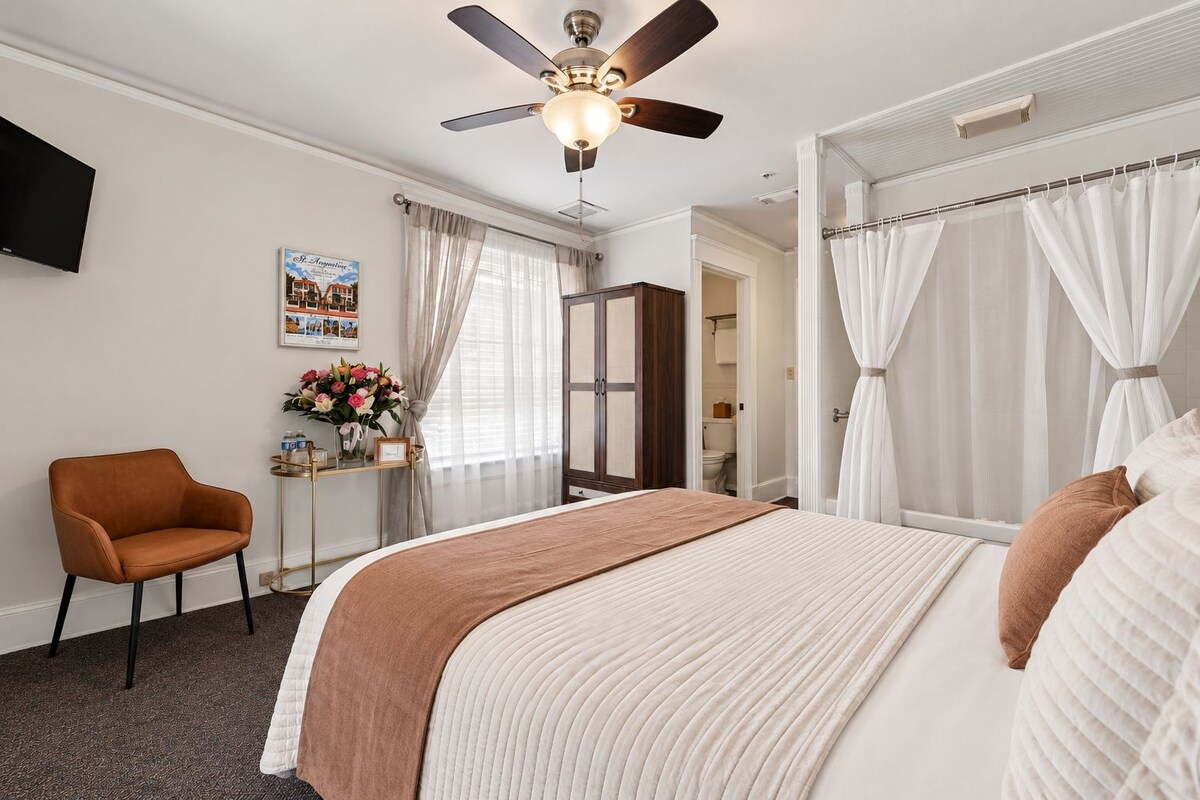 A hotel room with a bed, a chair, a desk with a vase of flowers, a ceiling fan, and a television on the wall.