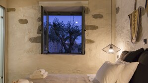 Un dormitori amb paret de pedra, una finestra amb vista a un arbre i a la lluna, un llit amb roba de llit blanca i una làmpada penjant.