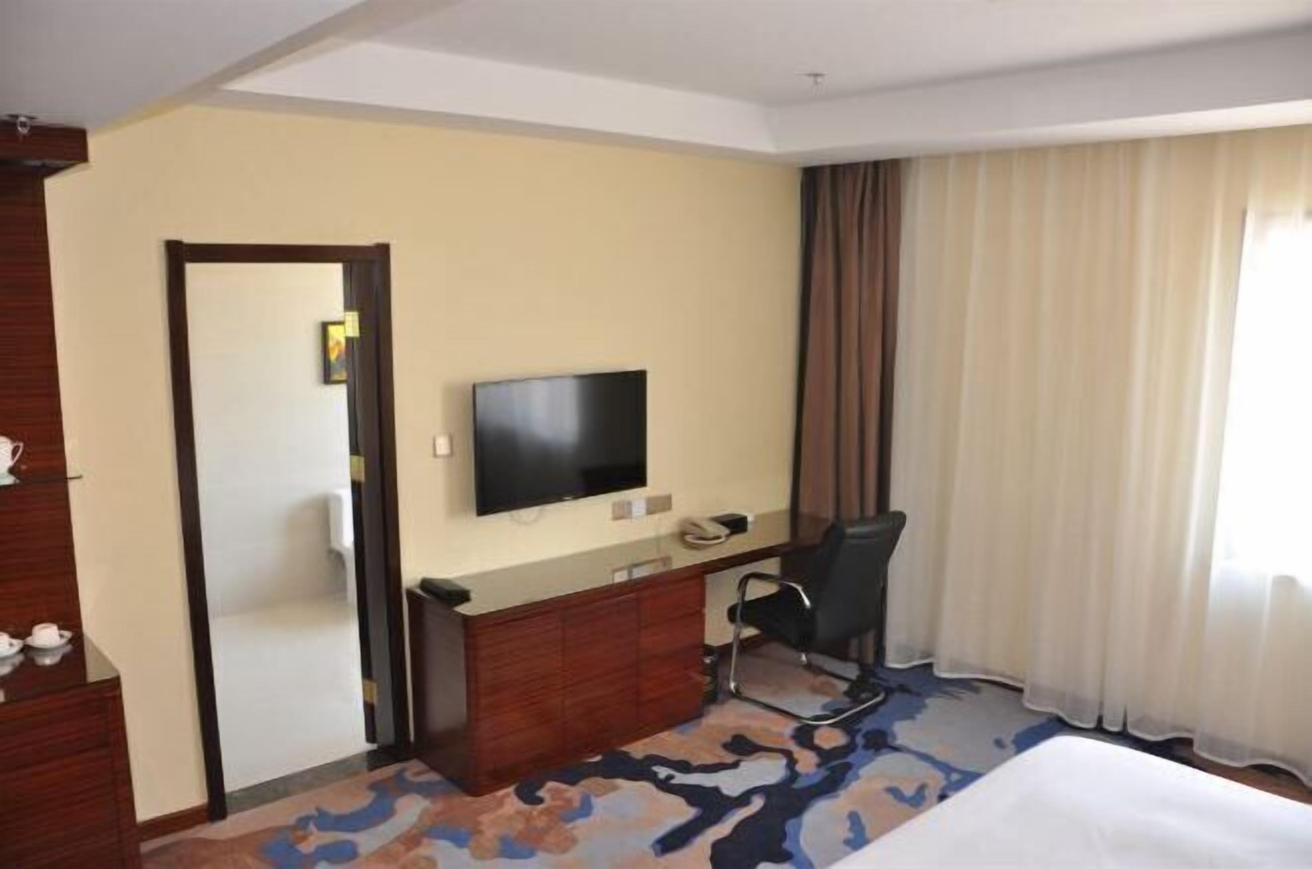 A hotel room with a bed, a desk with a chair, a television, and a large window with curtains.