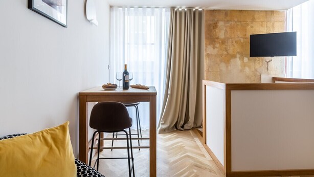 A hotel room with a wooden desk, a chair, a bed with a yellow pillow, a television on the wall, and a window with curtains. - Magnificent Bordeaux studio in beautiful stone in the heart of the city (Bordeaux)