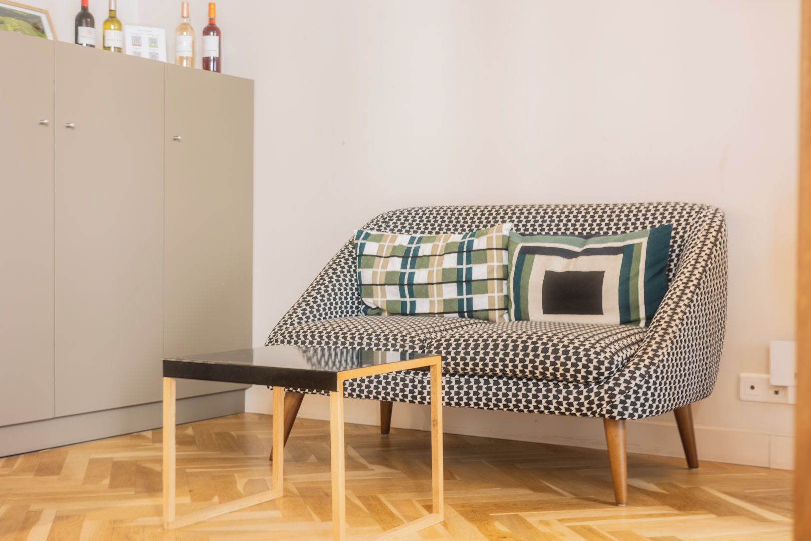 A houndstooth patterned armchair with a wooden frame and a matching ottoman, positioned in front of a cabinet with wine bottles.