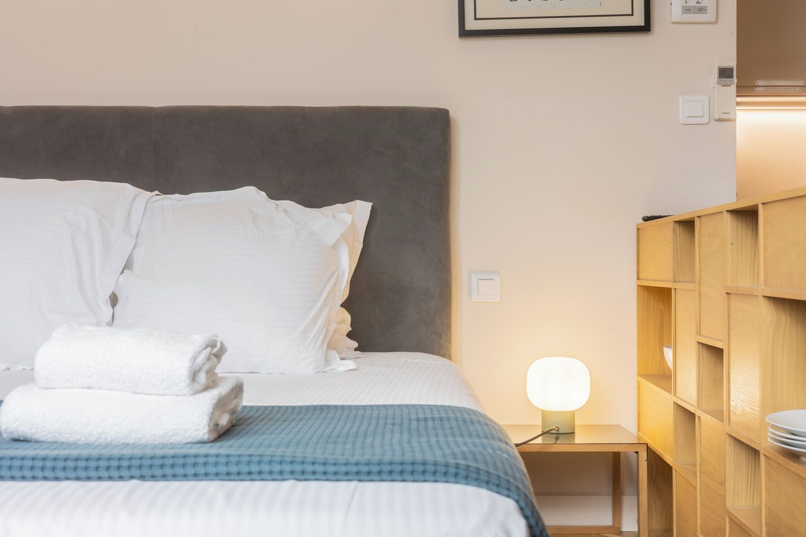 A neatly made bed with pillows and a folded towel, a bedside table with a lamp, and a wooden shelving unit.