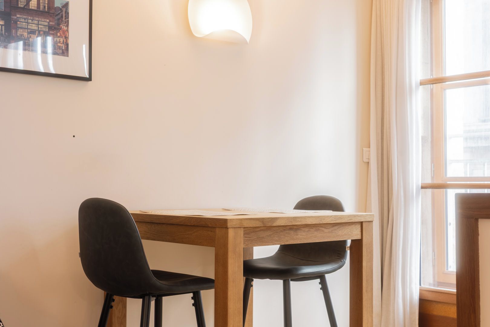 Quarto com uma mesa de madeira e duas cadeiras pretas, luminária na parede e uma janela com cortinas.
