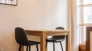 A room with a wooden table and two black chairs, a wall-mounted light fixture, and a window with curtains. - Magnificent Bordeaux studio in beautiful stone in the heart of the city (Bordeaux)