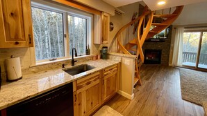 A kitchen with wooden cabinets, a granite countertop, a dishwasher, and a large window with a view of trees. - An Amazing Mountain Getaway! (Bartlett)