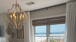 A dining area with a glass table, woven placemats, and wicker chairs. A chandelier hangs above, and a window offers a view of the beach. - OCEANFRONT 2 BD, 2.5 BTH in SURFSIDE! (Surfside Beach)
