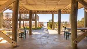 Un espace extérieur abrité, doté de piliers en bois, de tables de pique-nique et de chaises, qui mène à une terrasse en bois offrant une vue sur la plage.